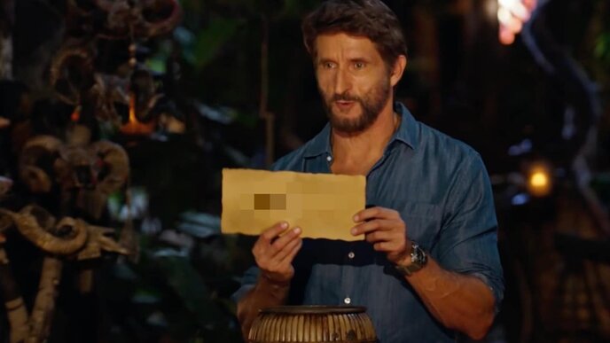 A man holding a pixelated letter in a jungle setting during a reality TV show, showcasing adventure, survival, and contestant drama on Reality Blurred