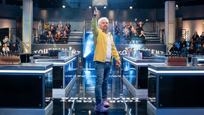 A man with white hair and a beard, wearing a yellow jacket, stands confidently on a game show set with multiple contestants and an audience in the background.