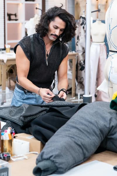 A man with curly black hair and a mustache, wearing a black sleeveless top, is working on a sewing project in a fashion design studio surrounded by mannequins and fabric.