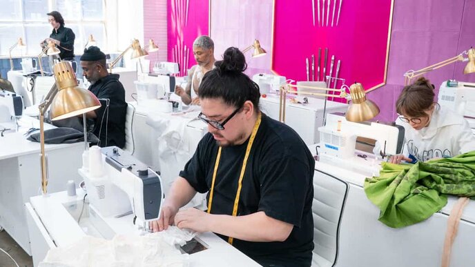 People working on sewing machines in a bright, modern workshop with pink and purple walls and gold lamps.